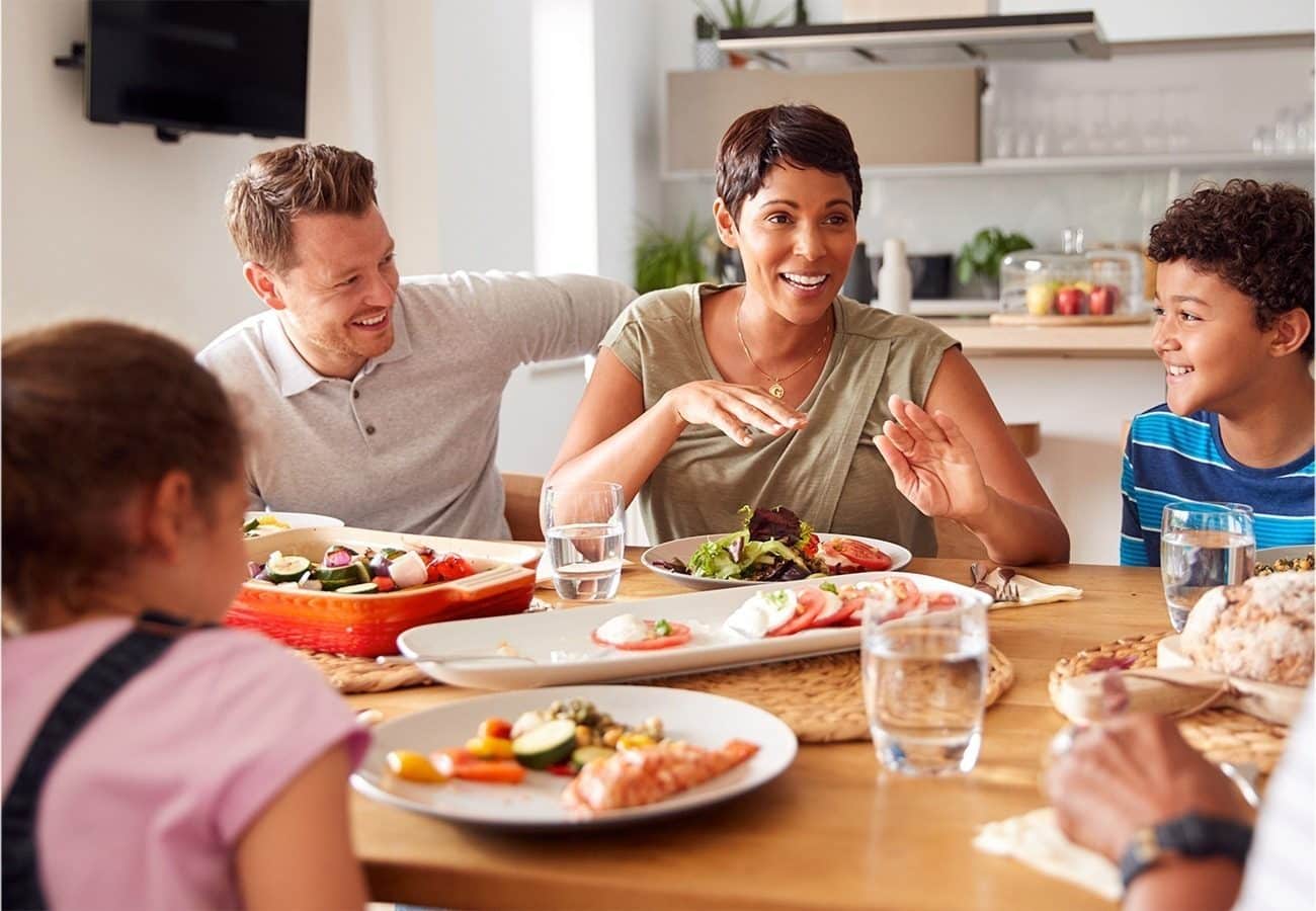 A family of four sits around a dinner table, smiling and talking while enjoying a meal together. Plates of food and glasses of water are on the table in a bright, modern dining area.
