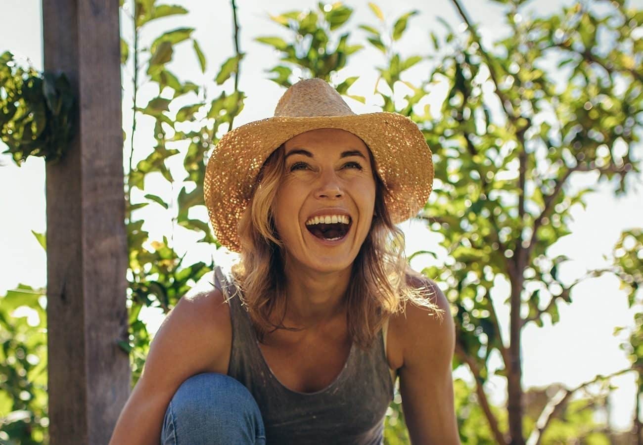 A woman wearing a straw hat and a sleeveless top laughs joyfully outdoors, surrounded by green leafy plants and sunlight.