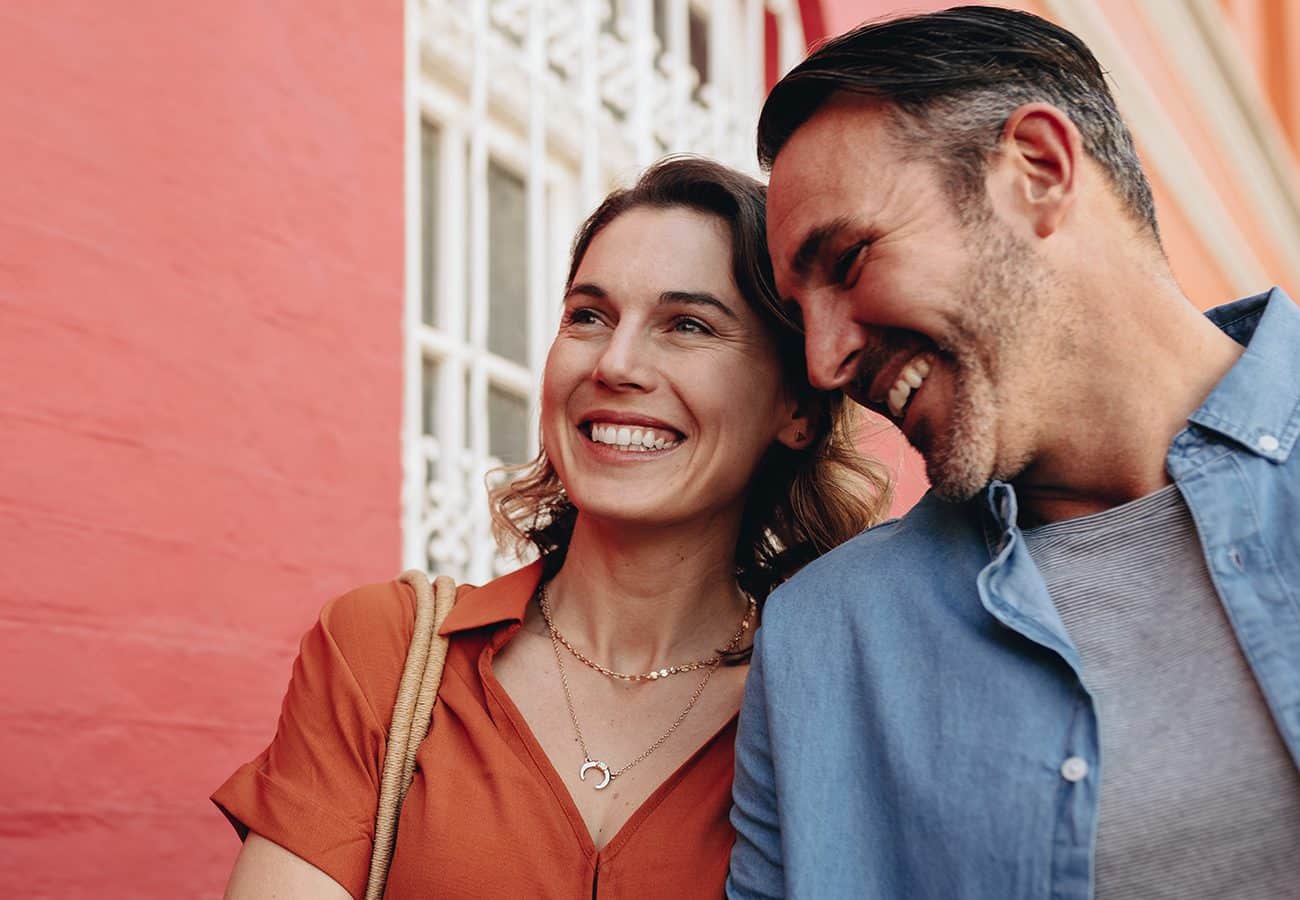 A woman in an orange shirt smiles while standing next to a man in a blue shirt who leans toward her, both appearing happy in front of a brightly colored wall with a white window grill.